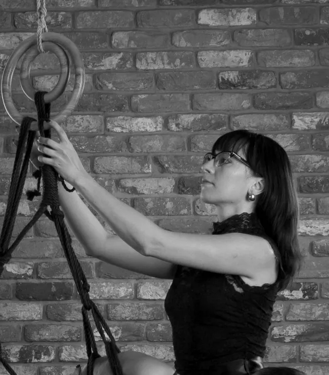 Yuliia Vasileva, in glasses and a black dress, reaches up to a wooden suspension ring while tying. Black-and-white archive photograph.
