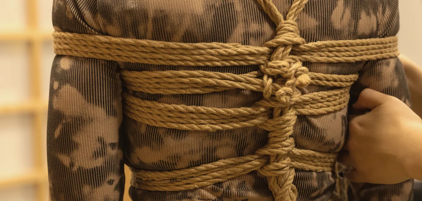 Close-up of a jute chest harness tied over a patterned top during a class at BONDAGE STUDIO; a teacher’s hand checks the tension.