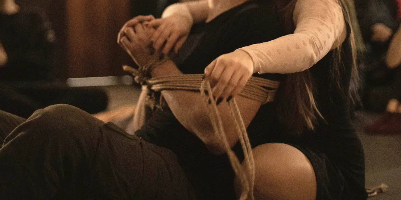 Warm evening light: one person gently binds another’s forearms with jute while seated on the floor.